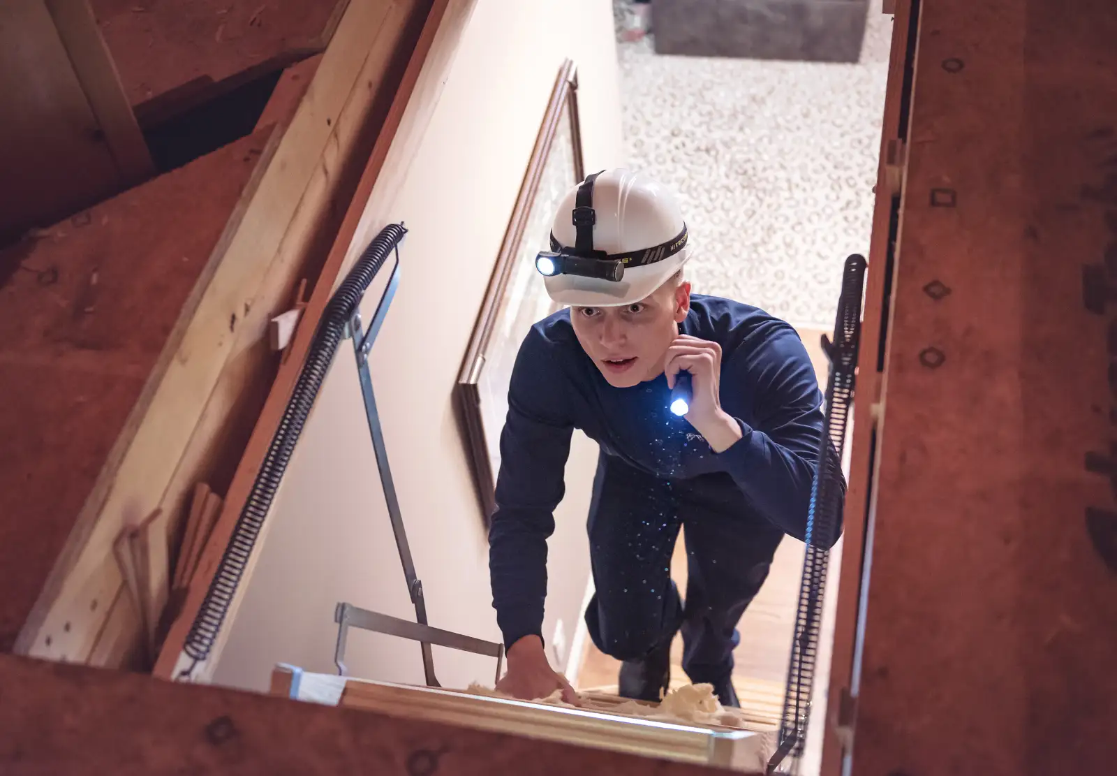 A Rocket Pest Control technician with a hard hat and headlamp climbs into an attic to inspect for pests.